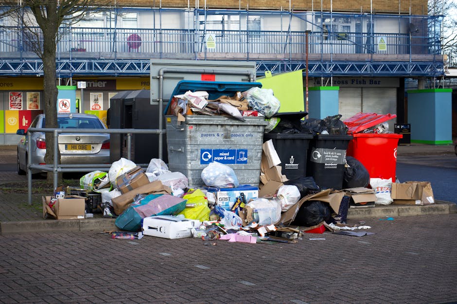 A worker wearing a yellow safety vest and black trousers is emptying waste into the rear compartment of a large red rubbish collection truck parked on the side of a street. The truck features various 