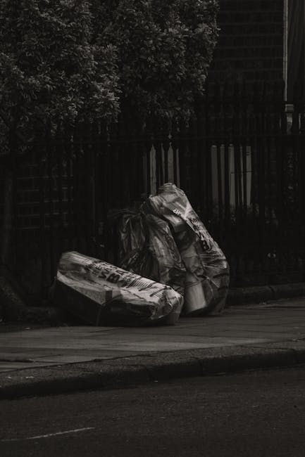 Three large black plastic rubbish bags filled with waste are positioned on a paved pavement beside a low curb. The bags are crumpled and appear to contain household or garden refuse. They are leaning 